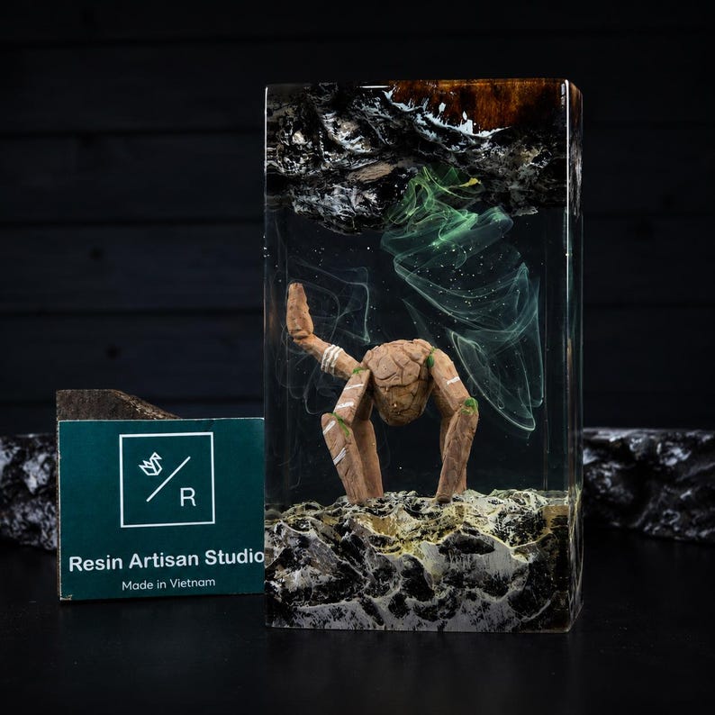 May include: A rectangular resin block featuring a miniature wooden creature with four legs standing on a textured base. The clear resin contains swirling green and white smoke-like patterns, with dark wood elements at the top and bottom edges.