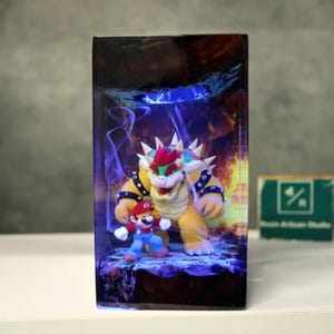 May include: A rectangular resin art piece featuring the characters Mario and Bowser from the Super Mario video game series. The figures are set against a backdrop of flames and blue lighting, with a dark wooden frame.