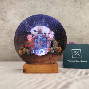 K&ouml;nnte beinhalten: Ein rundes Nachtlicht aus Harz mit Halloween-Motiv. Das Licht zeigt ein Spukhaus, einen Vollmond und drei Horrorfilmfiguren. Der Sockel ist aus Holz und hat eine warme braune Farbe.