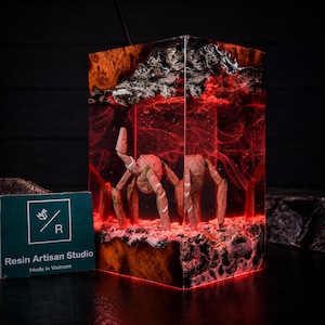 May include: A rectangular resin lamp featuring a dark wood base and top with a translucent red interior. Inside the resin, a light-coloured, multi-legged creature stands on a textured surface. A green card with the text Resin Artisan Studio is visible.