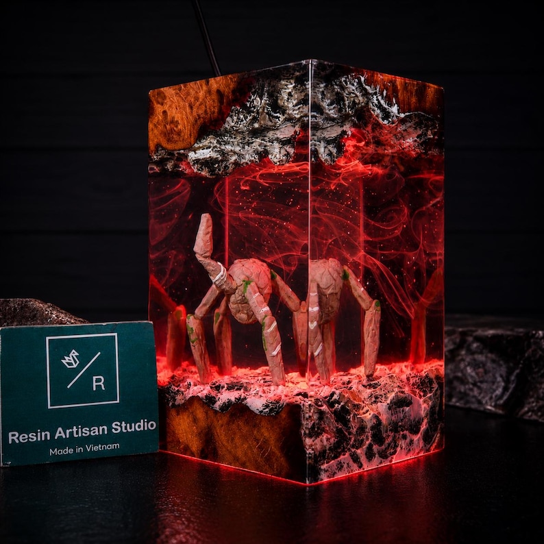 May include: A rectangular resin lamp featuring a dark wood base and top with a translucent red interior. Inside the resin, a light-coloured, multi-legged creature stands on a textured surface. A green card with the text Resin Artisan Studio is visible.