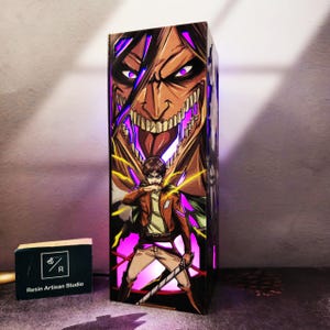 Könnte beinhalten: Rechteckige Kunstharzlampe mit einer Illustration aus der Anime-Serie Attack on Titan. Das Design zeigt das Gesicht eines Titans und eine Figur mit einem Schwert. Die Lampe hat ein leuchtendes Inneres mit violetten und gelben Akzenten. Auf der Basis steht "Resin Artisan Studio".