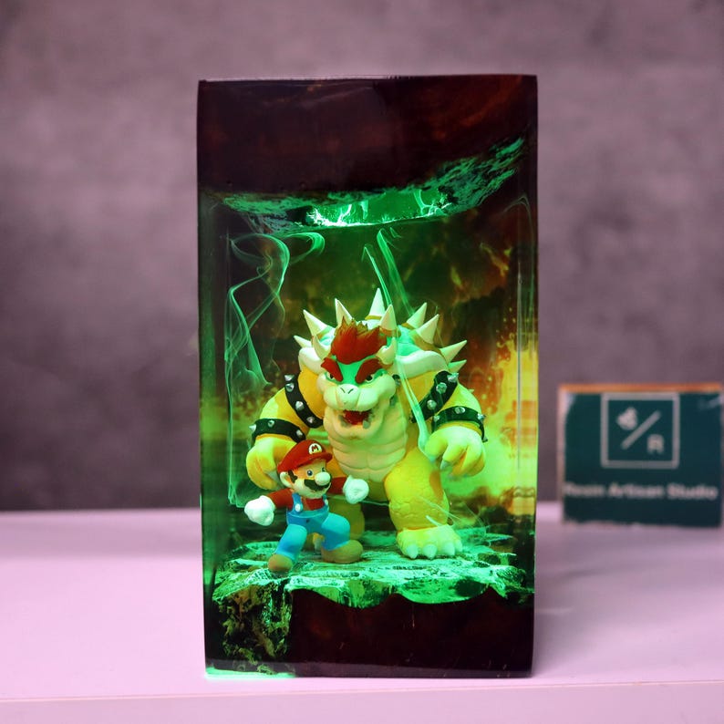 May include: A rectangular resin art piece featuring the characters Mario and Bowser from the Super Mario video game series. The figures are set against a green and yellow glowing background. The piece has a dark brown top and a small sign with text.