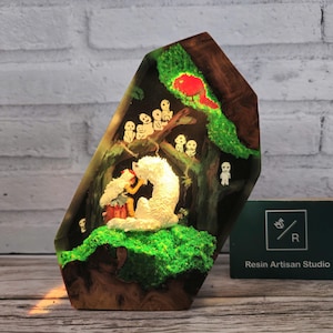 May include: A resin art piece depicting a scene from the movie "My Neighbor Totoro". A young girl is sitting with a white Totoro in a forest setting. The resin piece is set in a wooden base and has a green moss-like texture. The piece is illuminated from within, creating a warm glow.