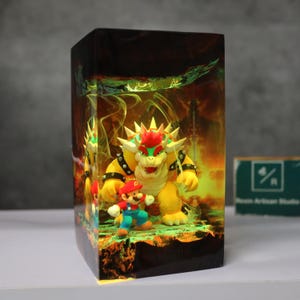 May include: A rectangular resin art piece featuring the characters Mario and Bowser from the Super Mario video game series. Bowser is yellow with red hair and spikes, and Mario is in his classic red and blue outfit. The background has a fiery, glowing effect.