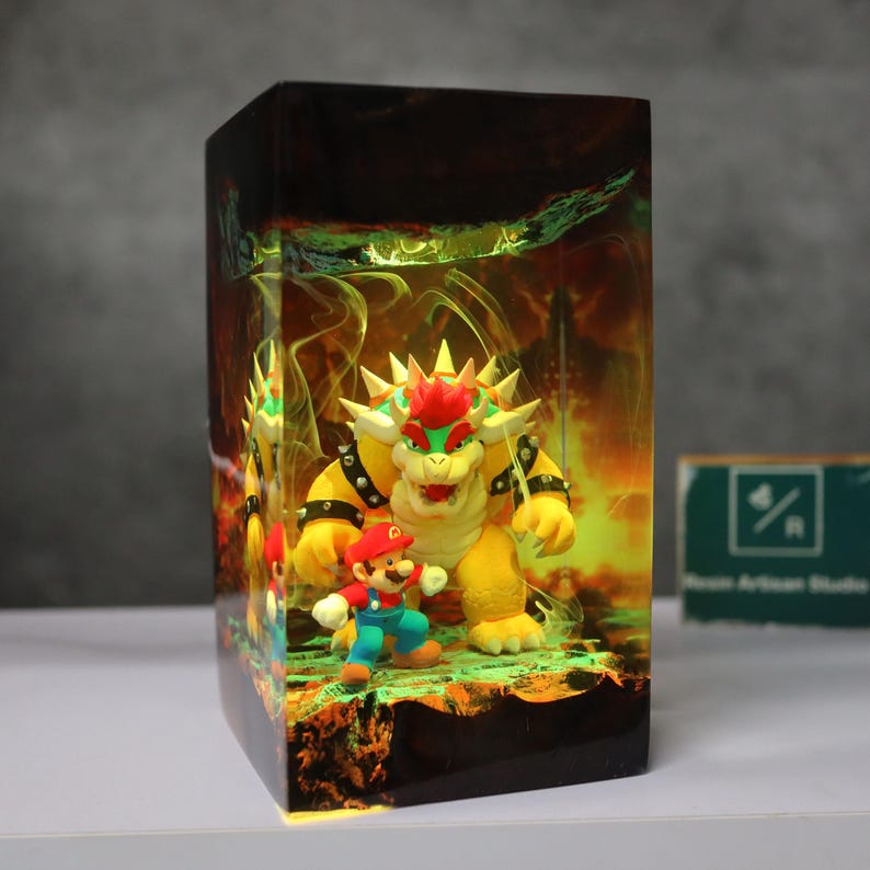 May include: A rectangular resin art piece featuring the characters Mario and Bowser from the Super Mario video game series. Bowser is yellow with red hair and spikes, and Mario is in his classic red and blue outfit. The background has a fiery, glowing effect.