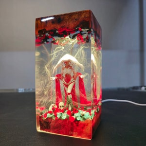 May include: A clear resin cube with a red and white figure inside. The figure is standing on a bed of red and green flowers. The cube is lit from inside, casting a warm glow on the figure and flowers.