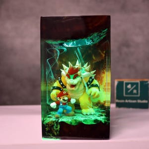 May include: A resin art piece featuring the characters Mario and Bowser from the Super Mario video game series. The figures are set against a green and yellow glowing background, encased in a rectangular block. The piece is on a pink surface.