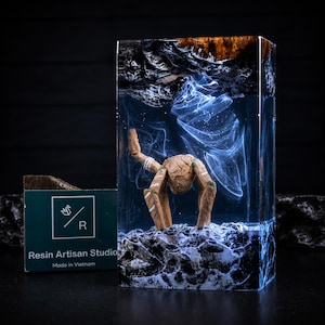 May include: A rectangular resin block featuring a suspended wooden creature with three legs and a textured surface. The clear resin contains swirling white smoke-like patterns above a dark, rocky base, with a dark wood top section.