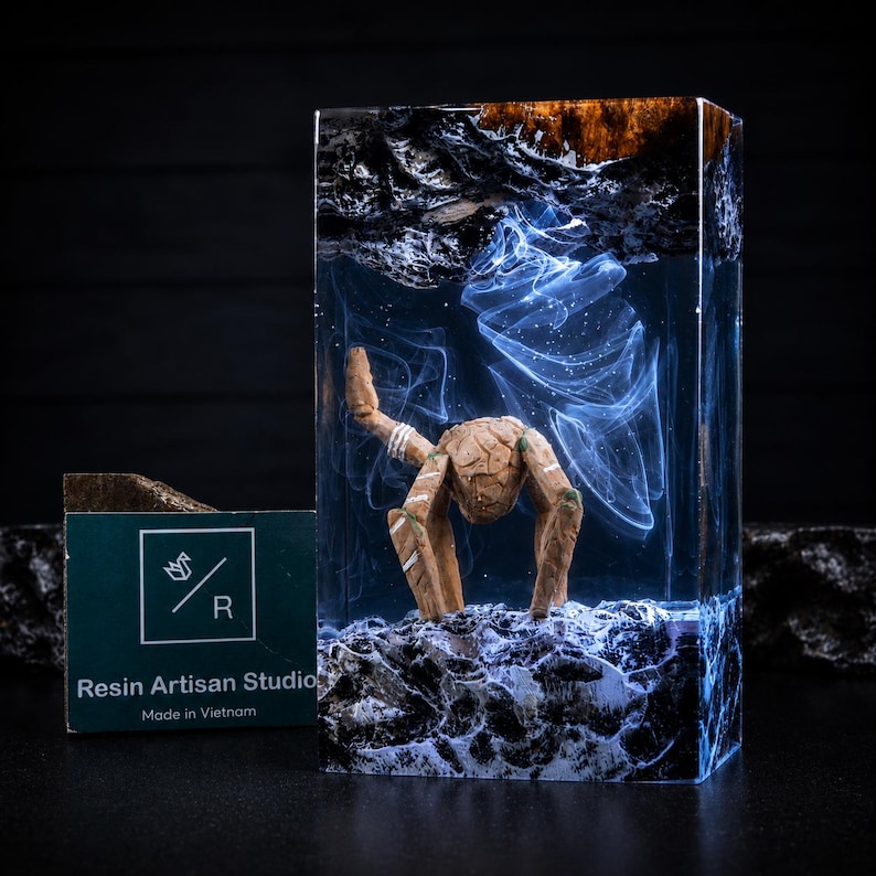 May include: A rectangular resin block featuring a suspended wooden creature with three legs and a textured surface. The clear resin contains swirling white smoke-like patterns above a dark, rocky base, with a dark wood top section.