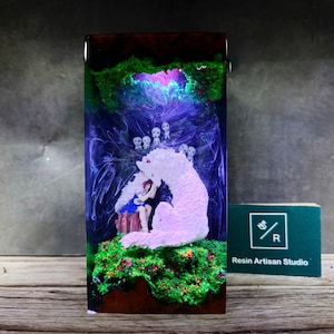 May include: A resin sculpture of a girl sitting with a white wolf in a forest setting. The sculpture is encased in clear resin with green, blue, and purple swirls. The wolf is glowing pink and the girl is wearing a blue dress. There are small white figures in the background.