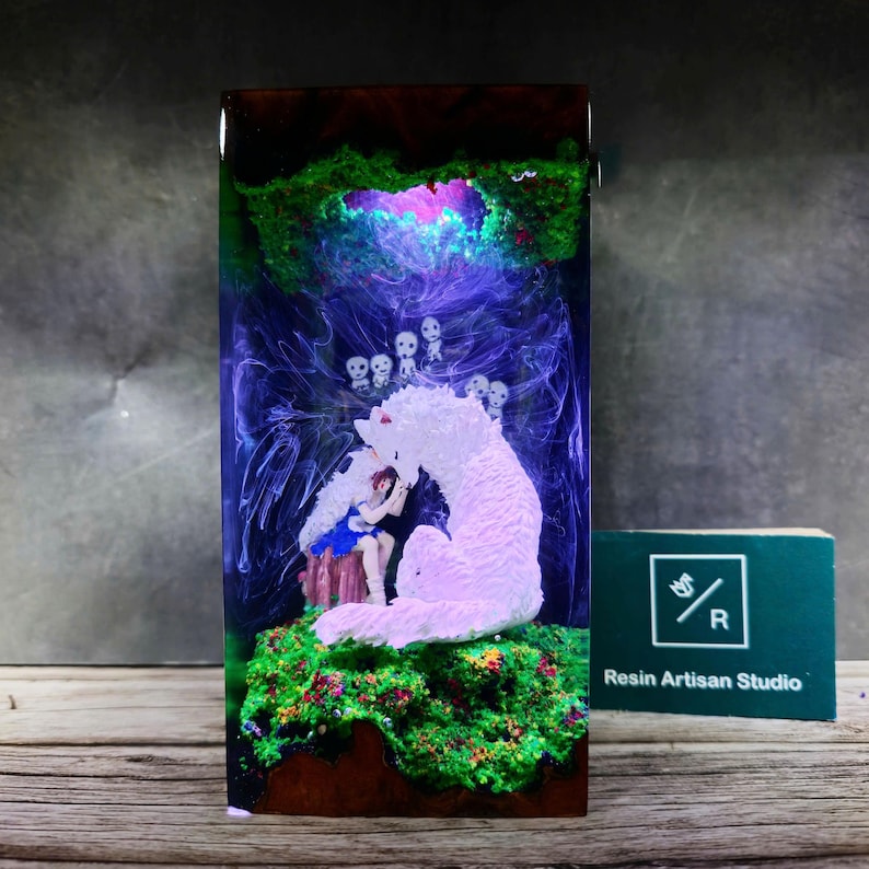 May include: A resin sculpture of a girl sitting with a white wolf in a forest setting. The sculpture is encased in clear resin with green, blue, and purple swirls. The wolf is glowing pink and the girl is wearing a blue dress. There are small white figures in the background.