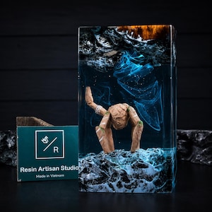 May include: A rectangular resin block featuring a submerged wooden figure resembling a creature with limbs, set against a dark blue swirling background. The top section has amber tones, while the base mimics rocky terrain. Text reads Resin Artisan Studio.