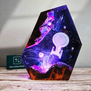 Puede incluir: Una pieza de arte en resina hexagonal que presenta un diseño de nave espacial sobre un vibrante fondo de galaxia. La obra de arte muestra una nave espacial blanca con detalles rojos y amarillos, rodeada de remolinos púrpuras, azules y blancos. La base tiene una textura similar a la madera.