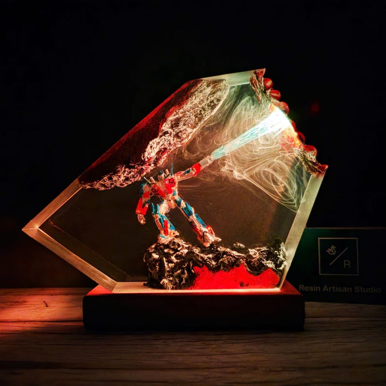 May include: A red, blue, and white robot figurine is encased in clear resin with a swirling blue light effect. The resin is set on a wooden base and is illuminated from within. The base has the text "Resin Artisan Studio" on it.