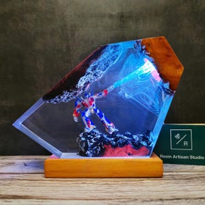 May include: A blue and red robot figurine is encased in clear resin with a blue and white swirl effect. The resin is set in a wooden base with the text "Resin Artisan Studio" on the front.