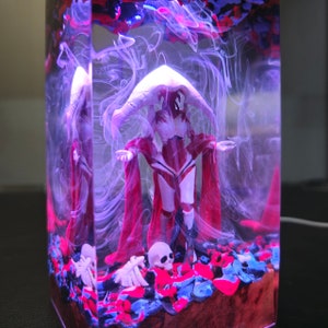May include: A resin sculpture of a figure wearing a red hooded cloak and white armor standing on a bed of red, blue, and black stones. The figure is surrounded by white smoke.