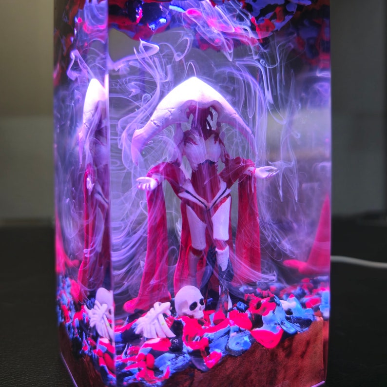 May include: A resin sculpture of a figure wearing a red hooded cloak and white armor standing on a bed of red, blue, and black stones. The figure is surrounded by white smoke.