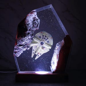 Könnte beinhalten: Eine Star Wars-Lampe mit dem Modell des Millennium Falcon, das in einem transparenten Harzblock aufgehängt ist. Die Lampe hat einen Holzsockel und enthält beleuchtete Details mit felsigen Formationen auf beiden Seiten des Schiffs. Der Hintergrund ist eine sternenklare Nacht.