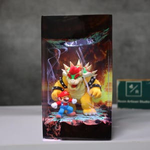May include: A rectangular resin diorama featuring Bowser and Mario figures. Bowser is yellow with a red mohawk, and Mario is in his classic red and blue outfit. The background depicts a fiery scene with smoke effects.