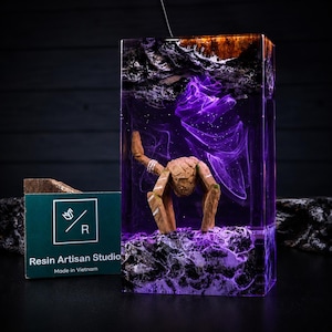 May include: A rectangular resin lamp featuring a dark wood top and base with a purple swirling smoke effect inside. A small, brown, articulated wooden figure stands in the centre. A green card with the text Resin Artisan Studio Made in Vietnam.