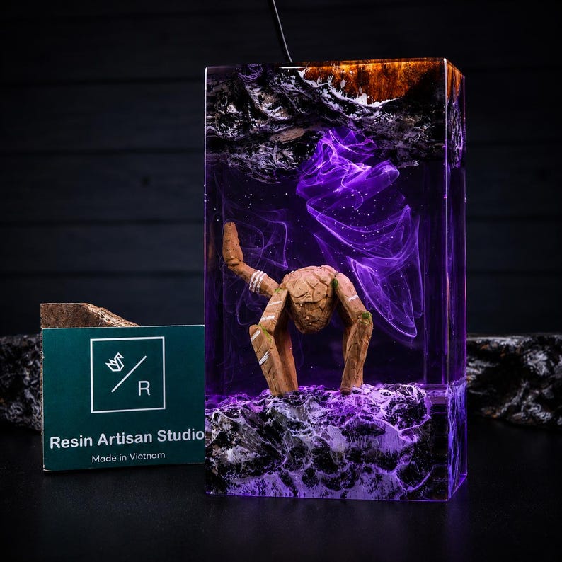 May include: A rectangular resin lamp featuring a dark wood top and base with a purple swirling smoke effect inside. A small, brown, articulated wooden figure stands in the centre. A green card with the text Resin Artisan Studio Made in Vietnam.