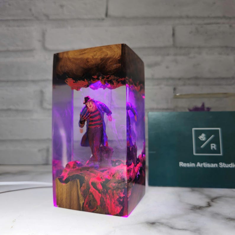 K&ouml;nnte beinhalten: Eine Harzlampe mit einer Freddy Krueger Figurine im Inneren. Die Lampe wird mit einem violetten Licht beleuchtet, und das Harz ist klar mit roten und schwarzen Akzenten. Die Lampe steht auf einer wei&szlig;en Oberfl&auml;che.