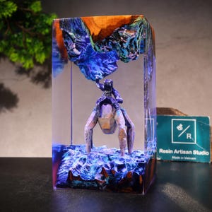 May include: A rectangular resin block containing a wooden landscape with a blue and purple glow. A small figure sits atop a four-legged creature beneath a jagged wooden overhang. A teal card with the text Resin Artisan Studio is visible nearby.