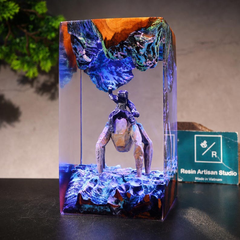 May include: A rectangular resin block containing a wooden landscape with a blue and purple glow. A small figure sits atop a four-legged creature beneath a jagged wooden overhang. A teal card with the text Resin Artisan Studio is visible nearby.