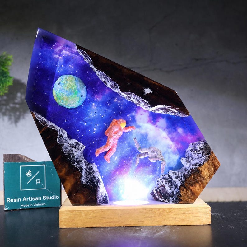 May include: A decorative resin lamp featuring a space scene with a blue and purple nebula, a green planet, and two floating astronauts. The piece is encased in a jagged, dark wood frame with white resin accents and sits on a rectangular wood base.