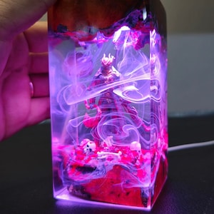 May include: A clear resin block with a red and purple glow. Inside the block is a small figurine of a demon with a skull and bones at its feet. The block is lit from the bottom, creating a dramatic effect.