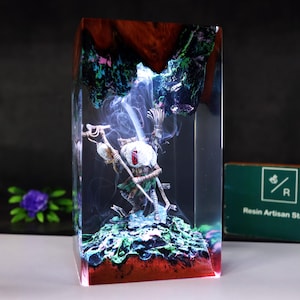 May include: A rectangular resin art piece featuring a skeletal figure with a staff, set within a clear block. The sculpture is surrounded by a smoky effect and rests on a green and blue base, with a red and brown top.