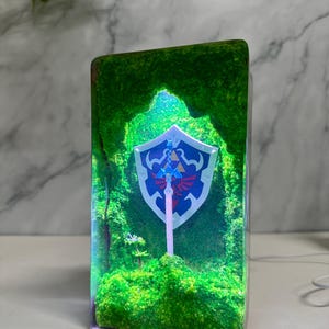 May include: A clear glass cube with a green moss base and a glowing blue and white sword and shield inside. The shield has a red and blue Triforce symbol.