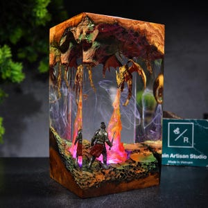 Może przedstawiać: Prostokątna lampa z żywicy i drewna przedstawiająca miniaturową scenę rycerza w srebrnej zbroi stojącego naprzeciw latającego smoka ziejącego pomarańczowo-żółtym ogniem. Podstawa i góra wykonane są z naturalnego drewna, wnętrze świeci.