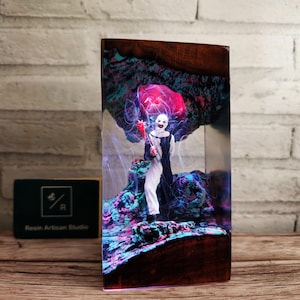 Op de afbeelding: Een harskunstwerk met een clownfiguur in een zwart-wit outfit, met een rood object. Het kunstwerk heeft een kleurrijke, getextureerde basis met een donkerhouten frame. Het stuk is verlicht met blauwe en roze verlichting.