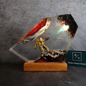 May include: A yellow robot figure with a blue energy beam shooting from its hand, encased in clear resin with a wooden base. The resin piece is shaped like a diamond and features a red and white rock formation.