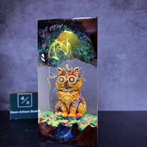 May include: A resin art piece featuring a regal cat figurine with a crown and goggles, set within a clear block. The cat is yellow, orange, and brown, with a colorful background. The base is a dark, textured surface.