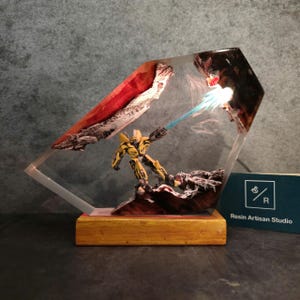 May include: A yellow robot figurine with a blue energy beam shooting from its hand, encased in clear resin with a red and white background. The resin piece is on a wooden base with a business card that reads "Resin Artisan Studio".