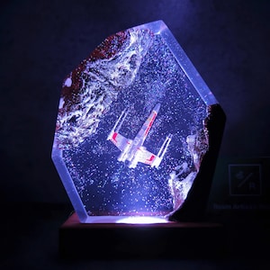 Puede incluir: Una lámpara de resina con una base transparente y brillante y una base de madera. Un caza estelar X-Wing en miniatura está incrustado en la resina, rodeado de purpurina. La lámpara está iluminada desde abajo, proyectando un brillo azul.