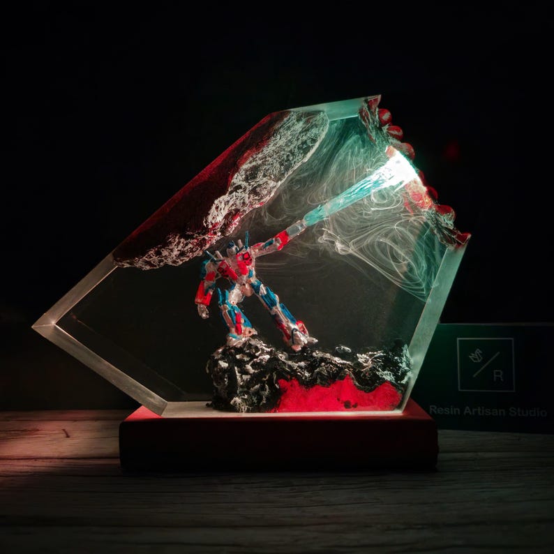 May include: A red, blue, and silver robot action figure is encased in clear resin with red and white swirling patterns. The resin is shaped like a diamond and has a light shining from the top. The base is red wood. "Resin Artisan Studio" is printed on a green card in the lower right corner.