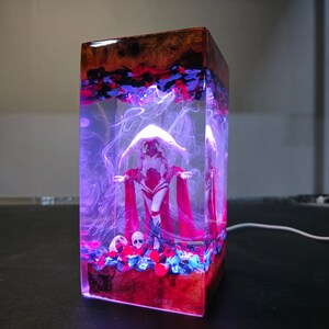 May include: A clear resin cube with a white figure wearing a red hooded cloak. The figure is standing on a bed of red, blue, and white stones. The resin cube is illuminated from within, casting a purple glow.
