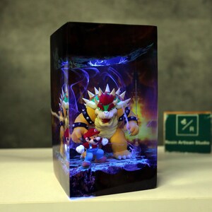 May include: A rectangular decorative box featuring a vibrant illustration of Bowser and Mario from the Super Mario video game series. The artwork includes a fiery background and blue energy effects. The box is on a white surface.