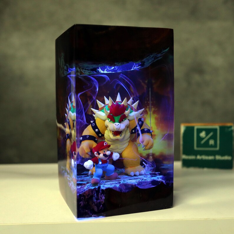 May include: A rectangular decorative box featuring a vibrant illustration of Bowser and Mario from the Super Mario video game series. The artwork includes a fiery background and blue energy effects. The box is on a white surface.