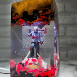 K&ouml;nnte beinhalten: Ein klarer Harzblock mit einem abstrakten Design in Rot, Schwarz und Gold. Im Inneren des Blocks befindet sich eine kleine Figur eines Horrorfilmcharakters, der eine Machete h&auml;lt. Der Charakter steht auf einem roten, schwarzen und wei&szlig;en Sockel.