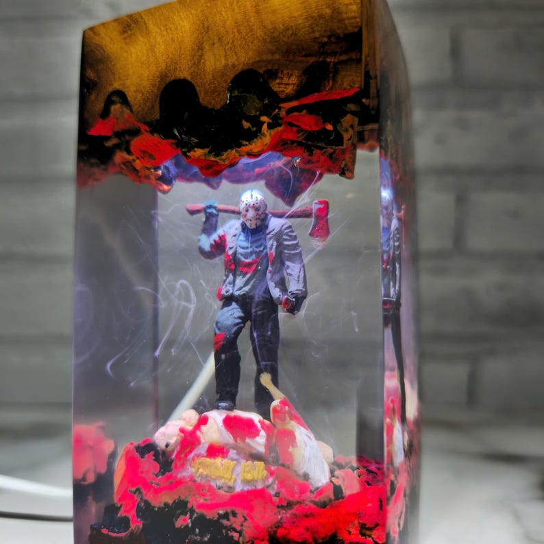 K&ouml;nnte beinhalten: Ein klarer Harzblock mit einem abstrakten Design in Rot, Schwarz und Gold. Im Inneren des Blocks befindet sich eine kleine Figur eines Horrorfilmcharakters, der eine Machete h&auml;lt. Der Charakter steht auf einem roten, schwarzen und wei&szlig;en Sockel.