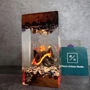 May include: A clear resin block with a small, black dragon figurine inside. The dragon is surrounded by orange and yellow resin, simulating fire. The block is sitting on a wooden base.