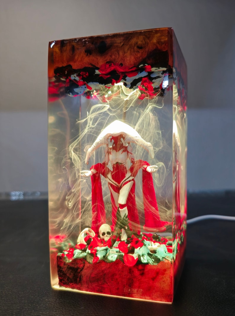 May include: A clear resin cube with a white figure wearing a red cloak and a white hood. The figure is standing on a bed of red flowers and a skull. The cube is illuminated from within, creating a glowing effect.