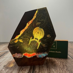 Puede incluir: Lámpara hexagonal de resina con diseño de la Enterprise de Star Trek. La lámpara tiene un fondo oscuro con detalles en rojo, amarillo y azul. La base es de madera oscura y la lámpara se ilumina desde dentro, creando un brillo cálido. Aproximadamente 15 cm de altura.