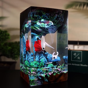 May include: A resin art piece featuring a detailed diorama of a fantasy scene. The artwork showcases figures with red, white, and blue accents, set against a backdrop of blue and green rock formations. The piece is encased in a clear rectangular block.