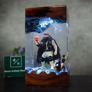 May include: A resin art piece featuring a figure holding a mushroom-shaped object, with a cat figurine. The artwork is encased in a clear resin block with a wooden base and top. The piece has blue and white accents, creating a unique display.